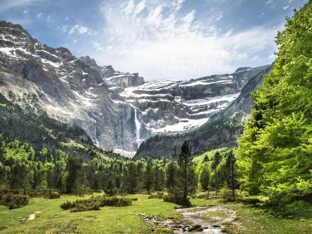 Vue d'une vallée verdoyante entourée de montagnes avec des glaciers au sommet et un ciel bleu.