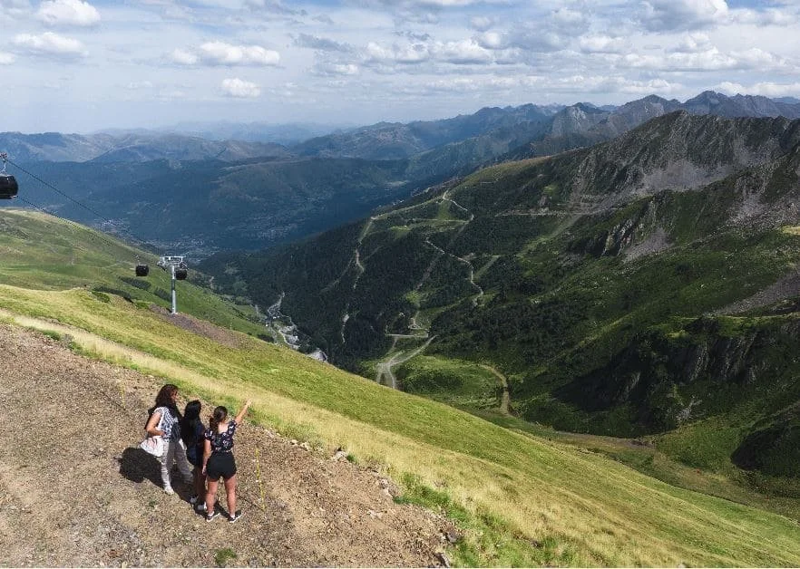 Trois femmes prennent une photo avec leur téléphone portable sur une colline verdoyante avec des montagnes en arrière-plan et un ciel partiellement nuageux.