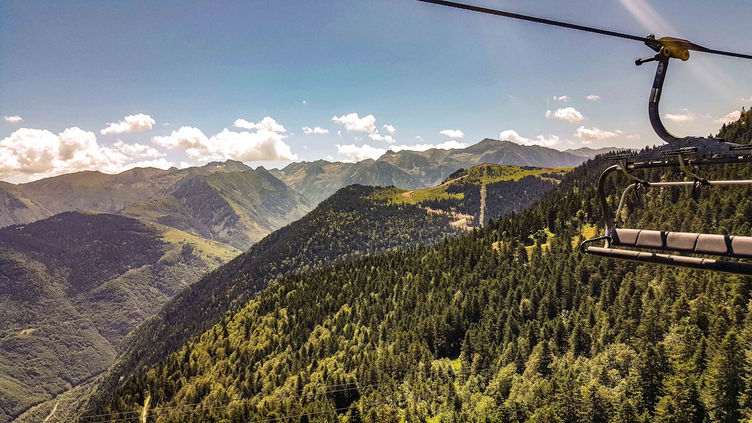 Vue de montagnes verdoyantes sous un ciel bleu avec quelques nuages, vue d'un téléphérique ou d'une chaise suspendue dans la partie droite de l'image.