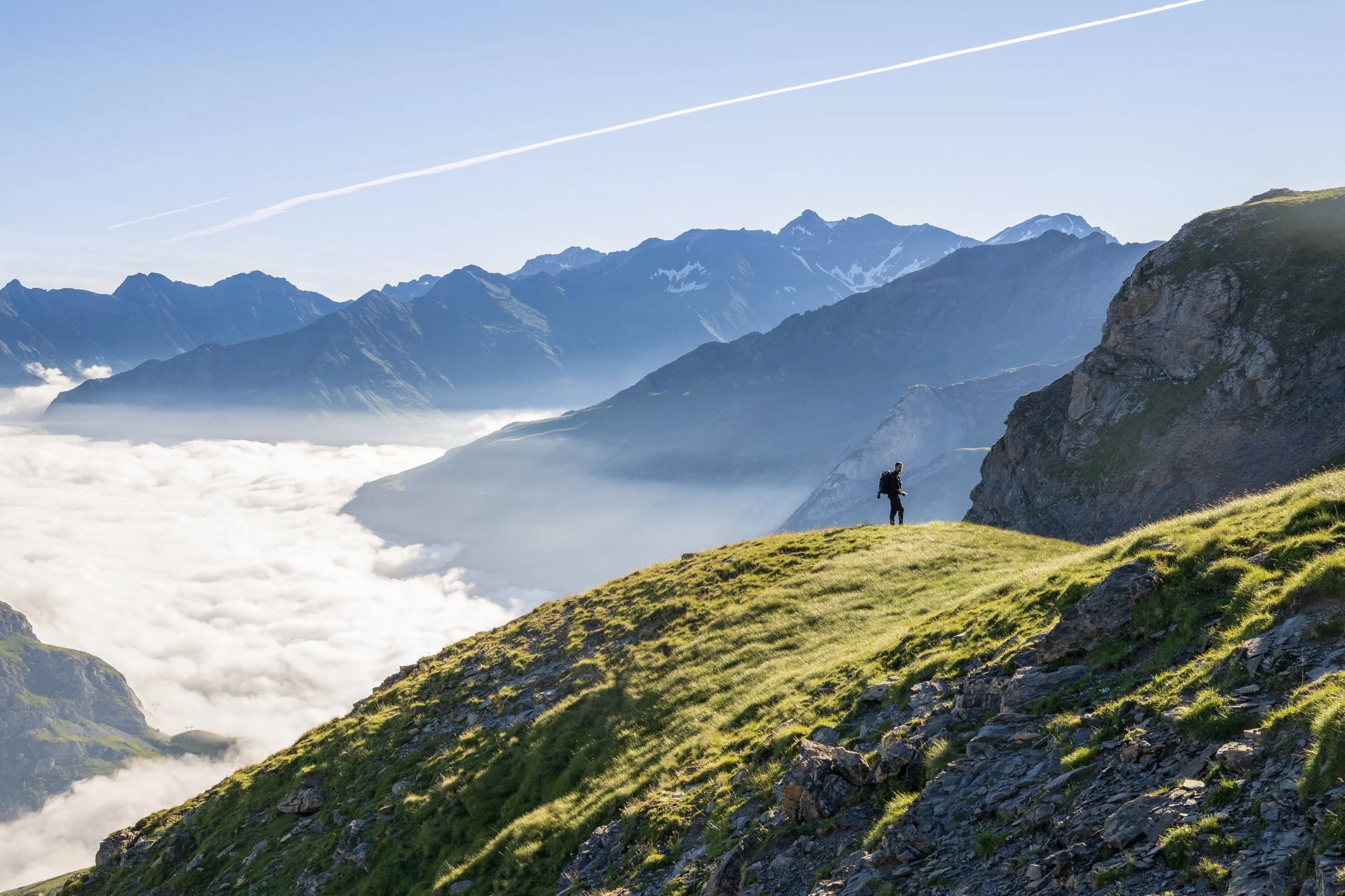 Un homme avec un sac à dos marchant sur une colline verte dans des montagnes, avec des nuages en dessous et un ciel clair.