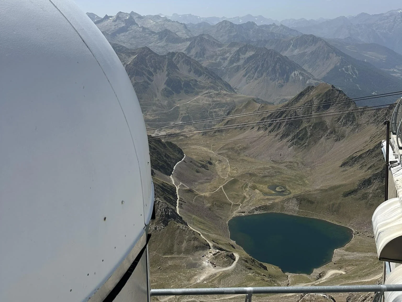 Vue aérienne d'une région montagneuse avec un lac au fond, des sentiers et un équipement de téléphérique en premier plan.
