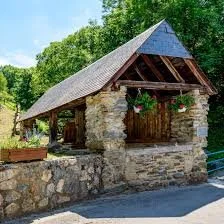 Une petite maison en pierre avec un toit en bois, entourée d'arbres verts, avec des fleurs suspendues à la façade.