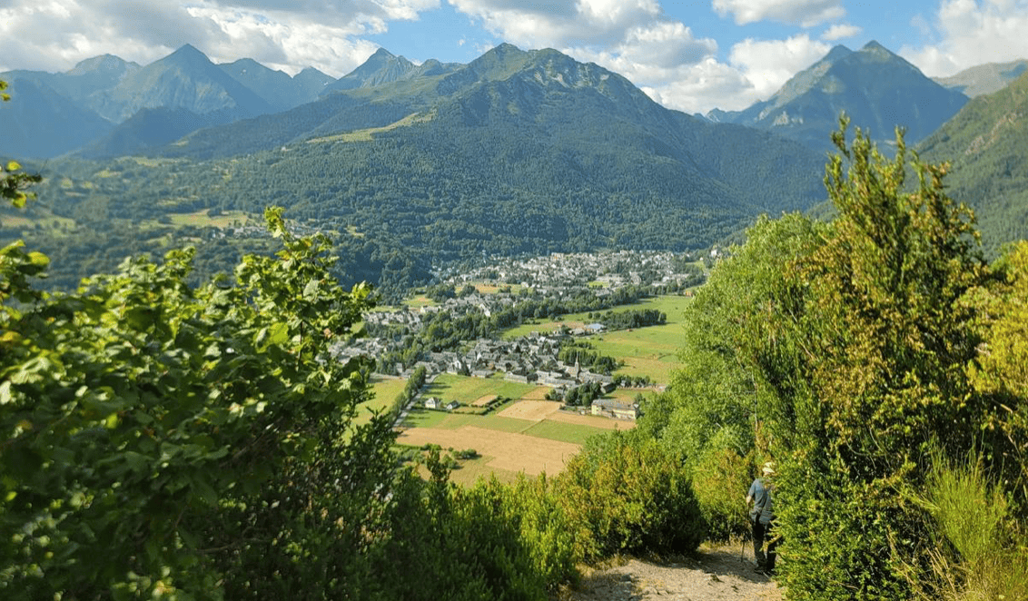 Paysage de montagnes verdoyantes avec un village dans la vallée, vu depuis un sentier entouré d'arbres, avec deux personnes marchant sur le sentier.