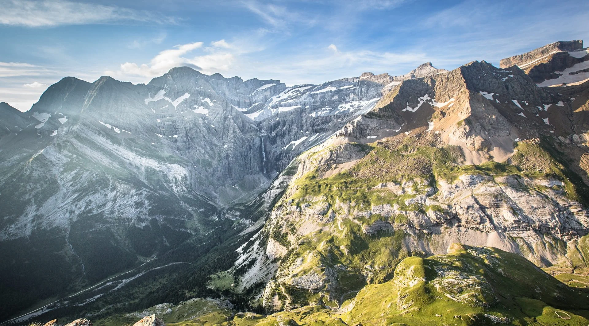 Montagnes rocheuses avec des glaciers, présence de neige, plaine verte en bas, ciel bleu avec quelques nuages