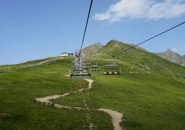Télésiège suspendu au-dessus d'une pente verdoyante dans un paysage de montagne avec des sommets en arrière-plan et un ciel partiellement nuageux.