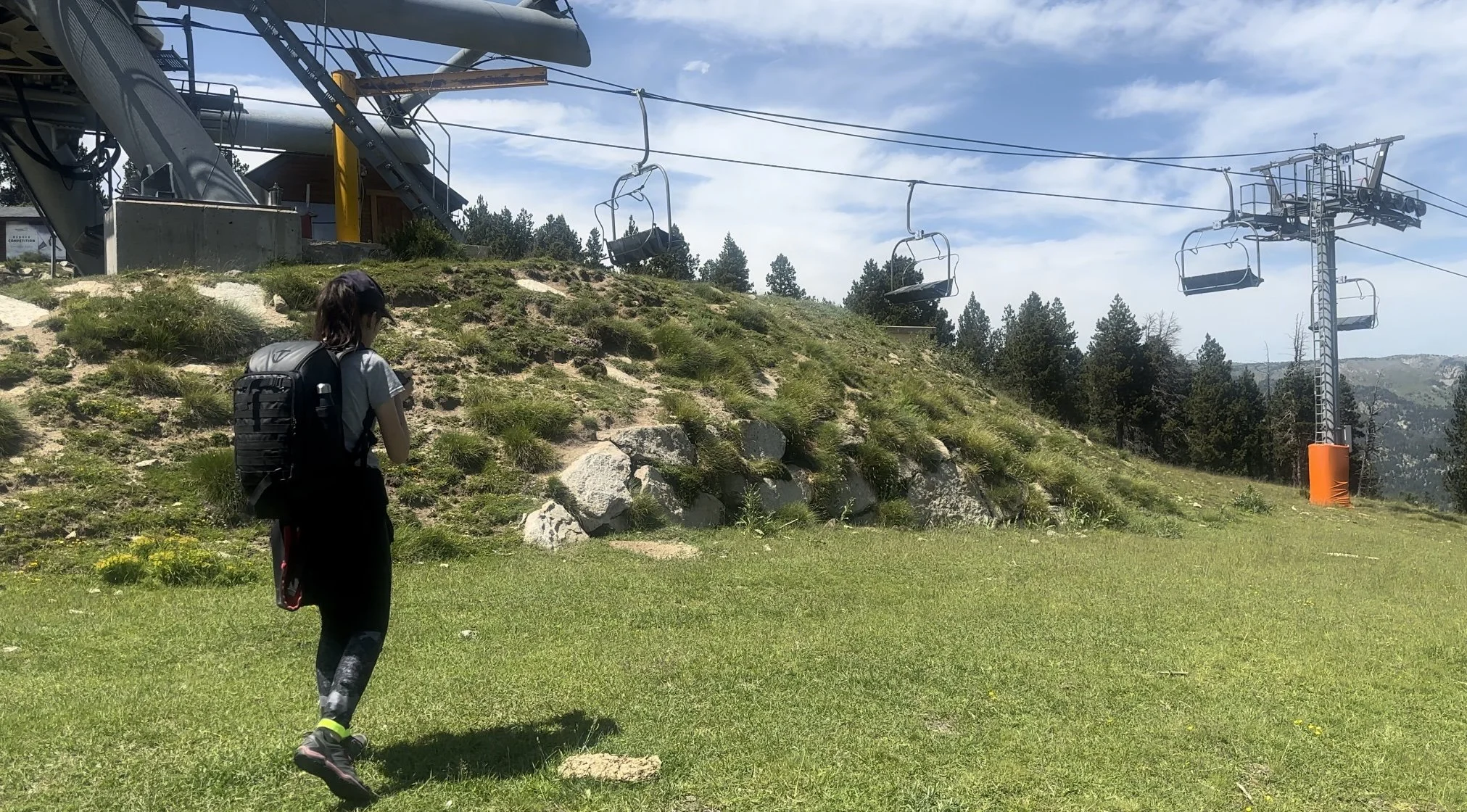 Une jeune fille avec un sac à dos regarde une télécabine en montagne, entourée de verdure et de arbres, ciel partiellement nuageux.