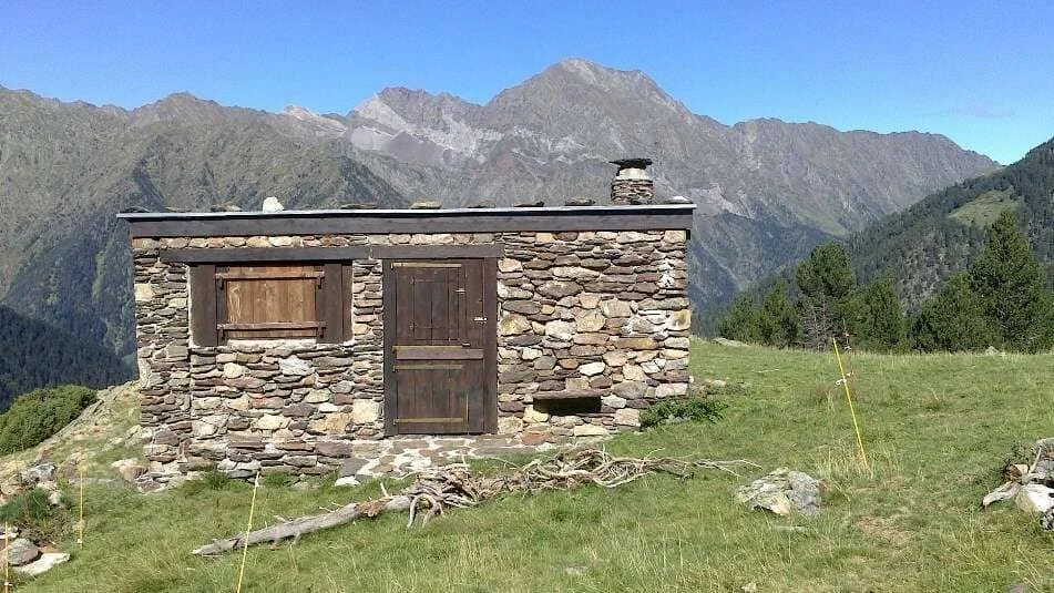 Une petite cabane en pierre au milieu d'une prairie avec des montagnes en arrière-plan.