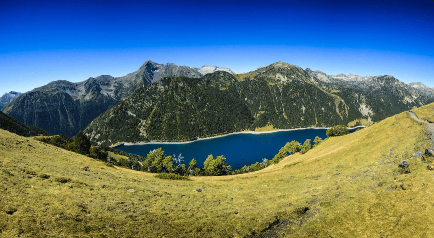Paysage de montagnes avec un lac en dessous, végétation verte et sommet rocheux sous un ciel bleu clair