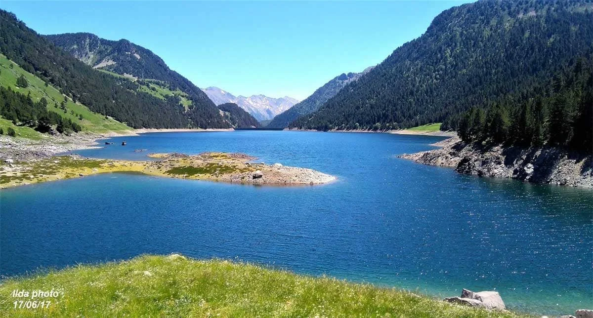 Lac entouré de montagnes verdoyantes sous ciel clair, avec un petit îlot au centre.