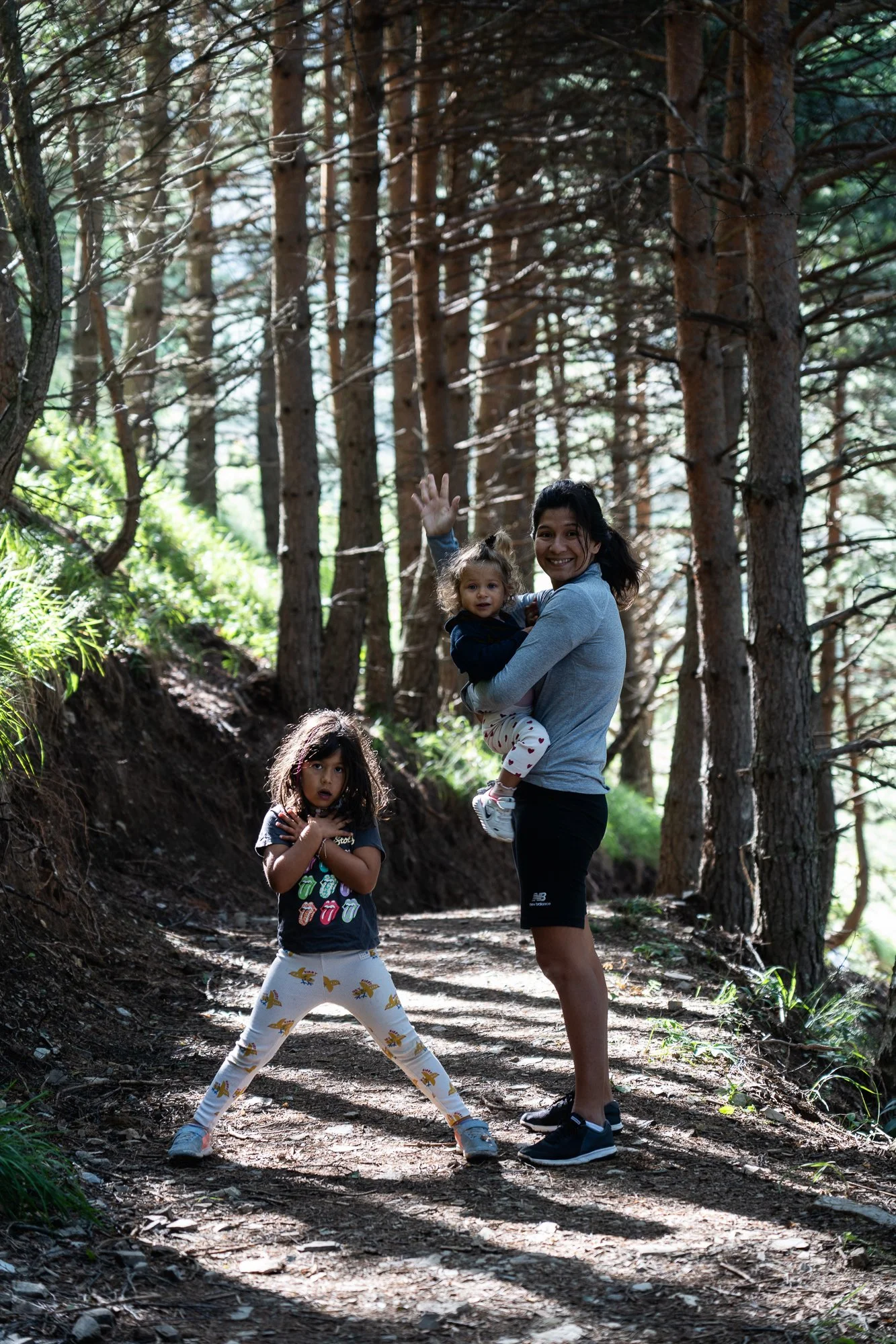 Une femme souriante tient dans ses bras une petite fille dans une forêt de pins. Il y a aussi une autre fille avec des cheveux bouclés, portant un t-shirt avec des lèvres en couleur et un pantalon blanc avec des motifs de dinosaures. Elle semble tris