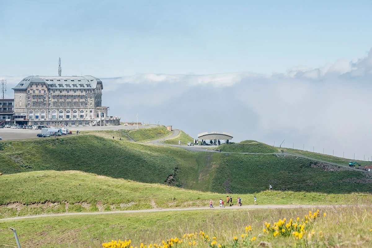 Paysage de collines vertes avec un grand bâtiment en pierre en fond, un parking, et des personnes marchant dans la nature, avec un ciel partiellement couvert de nuages.