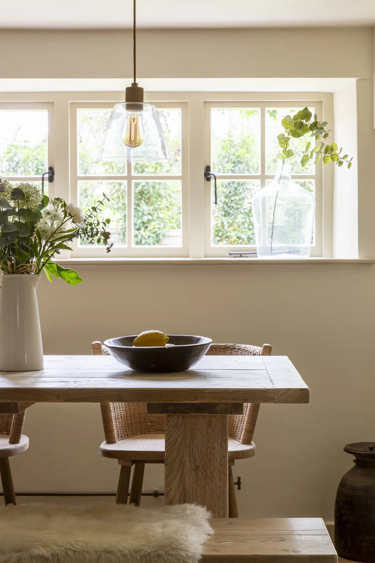 A cozy dining area with a wooden table, two wicker chairs, a black bowl with a lemon, a white vase with green and white flowers, a large glass vase with green branches on the window sill, and a pendant light hanging above the table. The window behind reveals greenery outside.