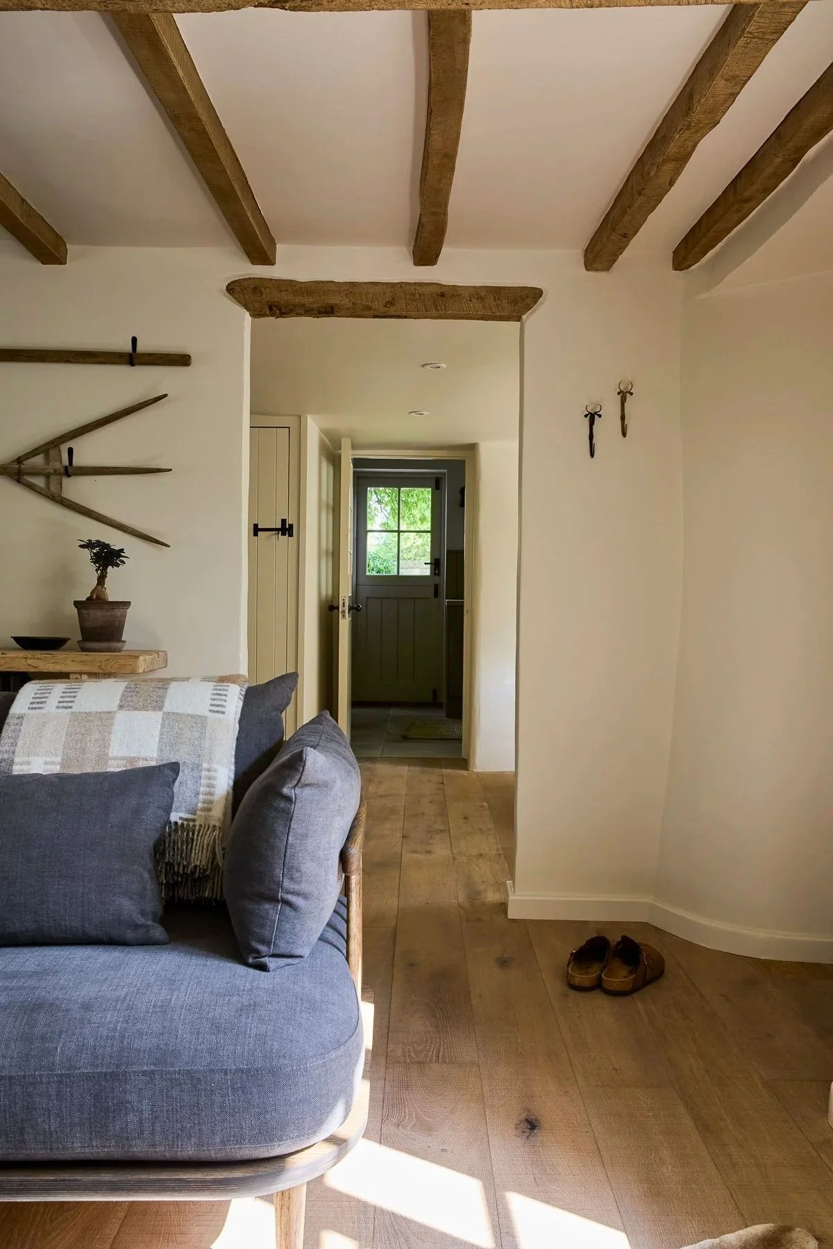 Cottage interior with exposed timber beams, warm neutral walls and a blue upholstered seat, looking through a doorway towards a glazed garden door.