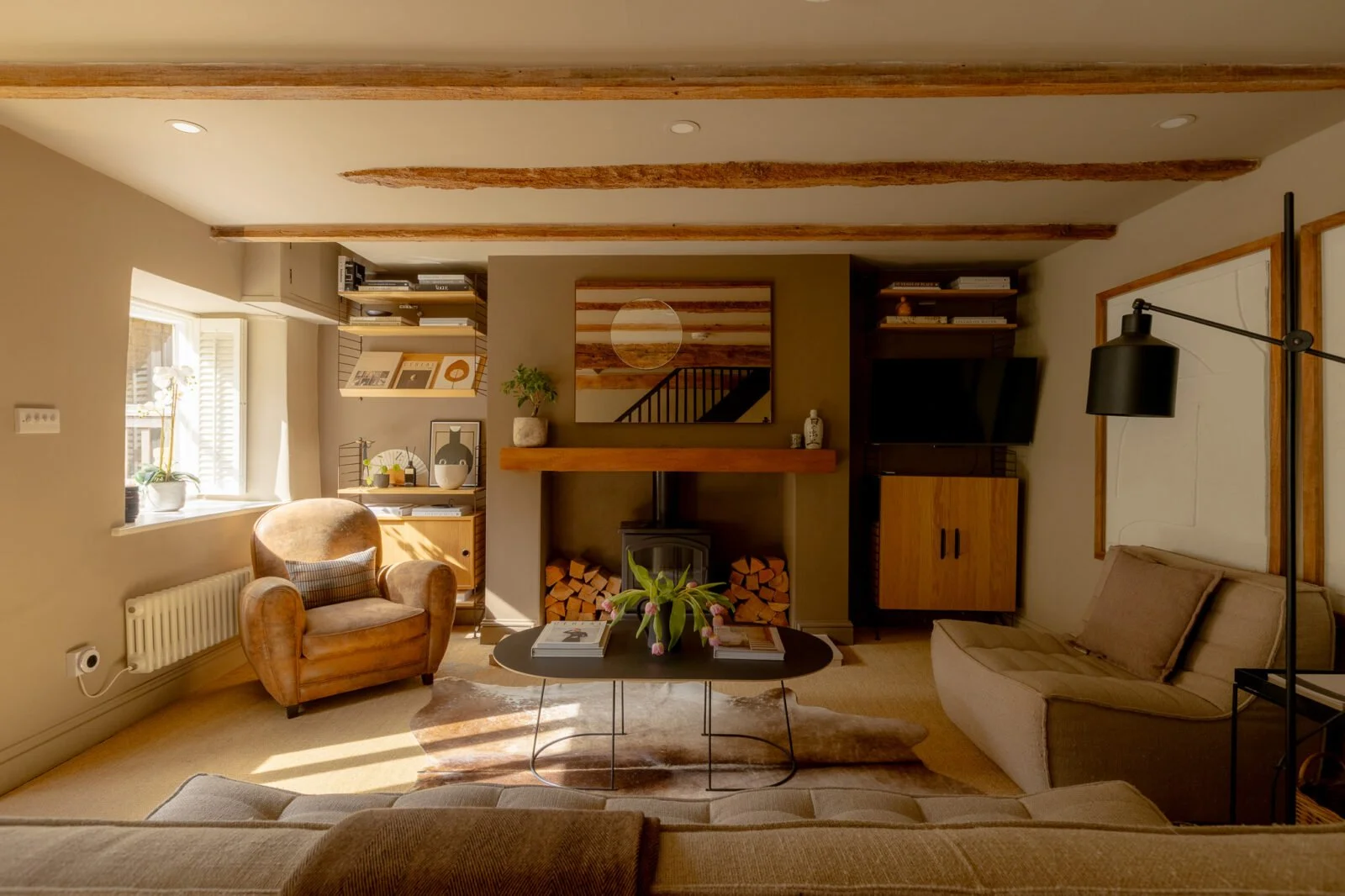 Period sitting room with exposed ceiling beams, a wood-burning stove, built-in shelving, a tan leather armchair and neutral sofas.