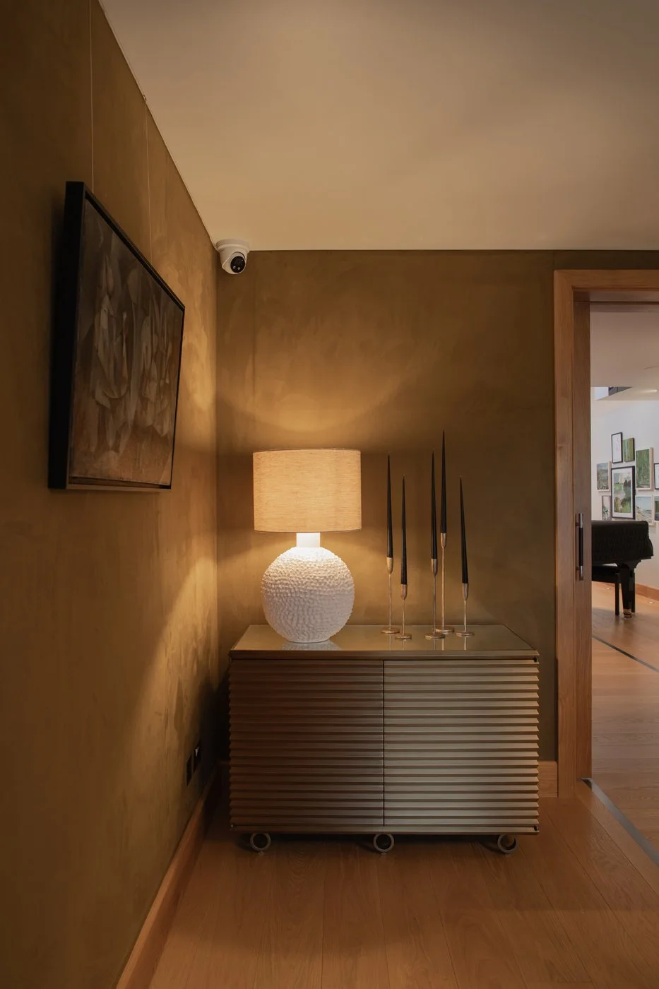 Textured white ceramic lamp and slender candlesticks on a console against warm bronze-toned walls, with artwork and a doorway beyond.
