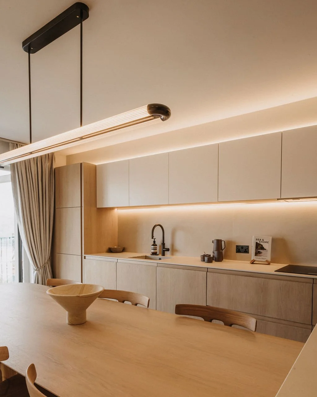 A warm, neutral-toned kitchen and dining area with pale wood cabinetry, soft under-cabinet lighting, a wooden dining table and a ceramic bowl centerpiece.