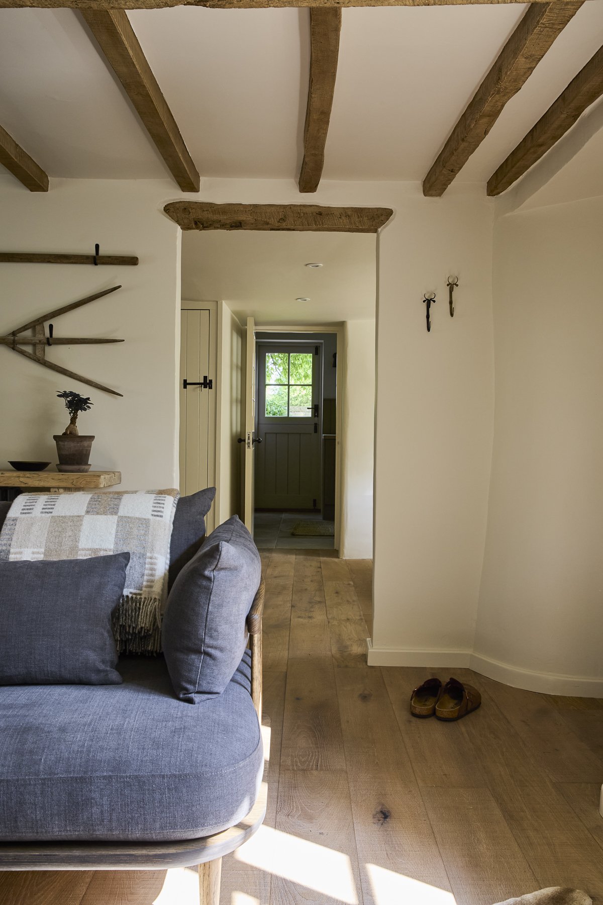 Interior view of a living room with wooden beams on the ceiling, a gray sofa with cushions, a wooden table with a potted plant, and a pair of shoes on the wooden floor. There's a doorway leading to an outdoor area visible through a windowed door.