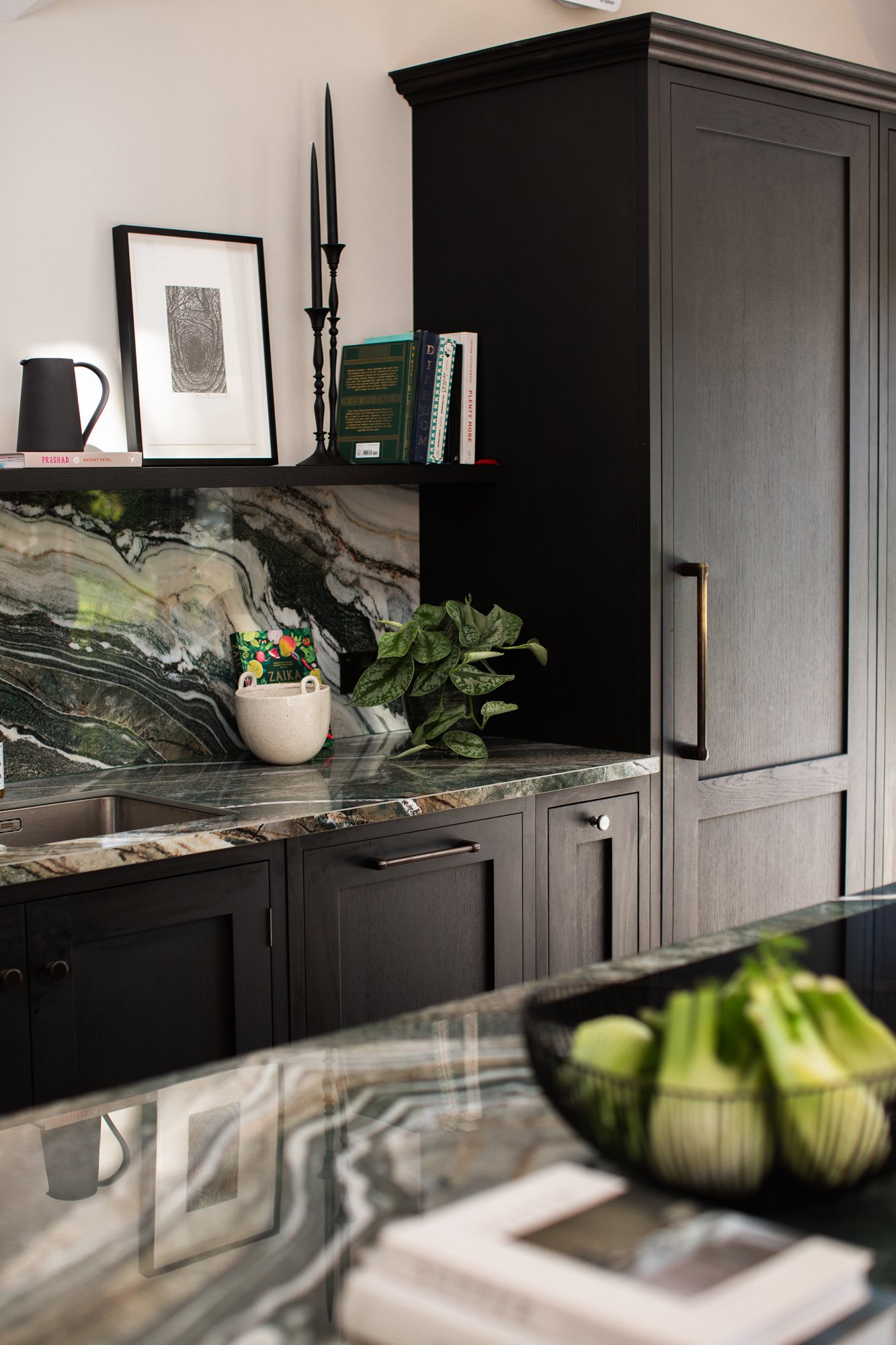 Refined kitchen detail with dark timber cabinetry, dramatic marble surface, books, ceramics and trailing greenery.