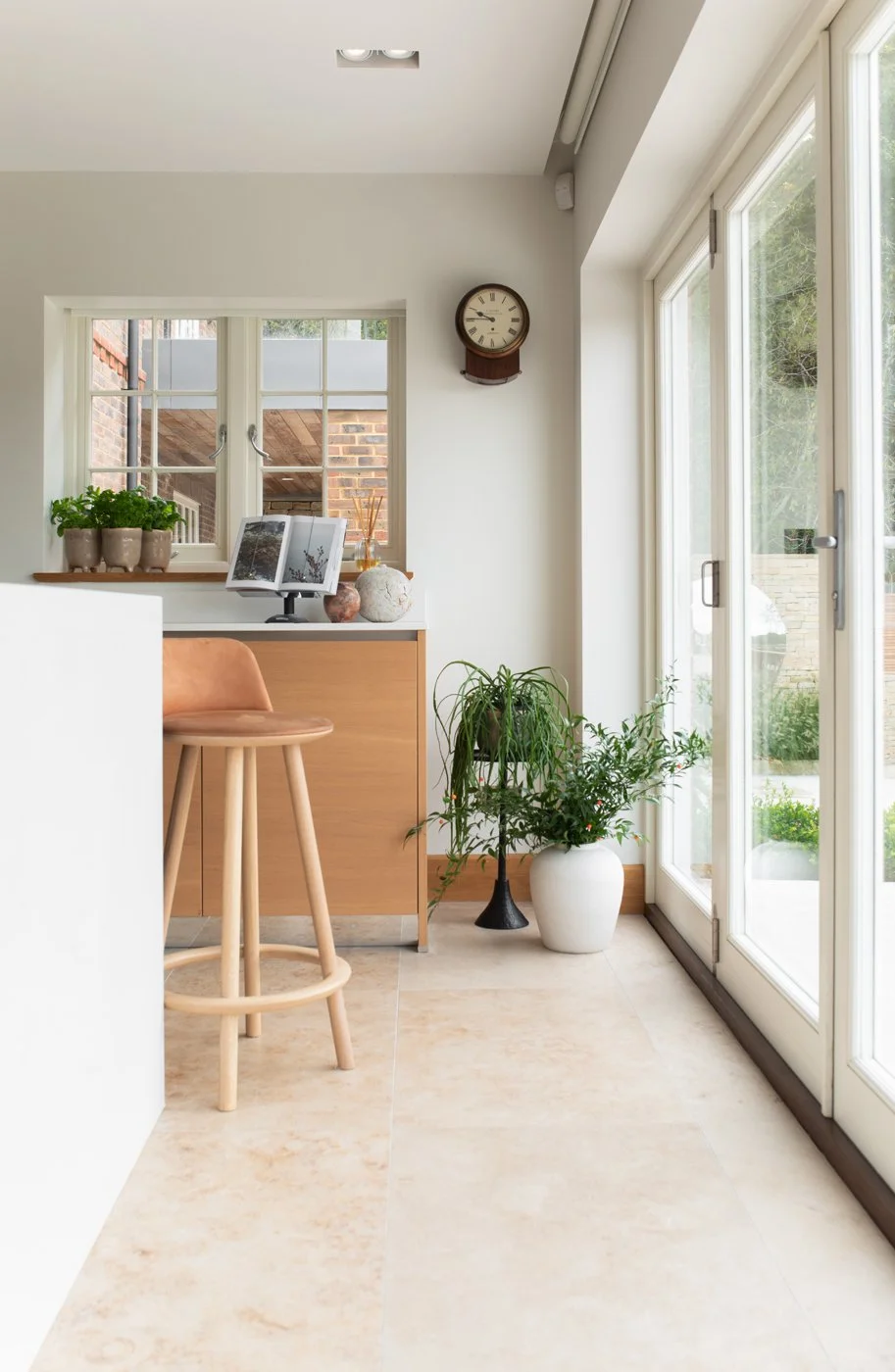 Bright indoor space with a large glass sliding door leading outside, with plants in white pots, a wooden stool, a wall clock, and decorative items on the window sill.