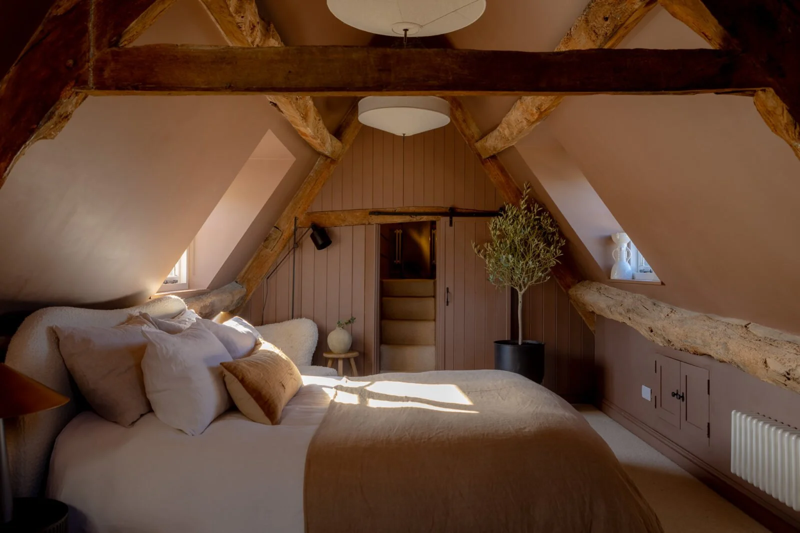 Bedroom at Bear Court Cottage in Burford, with exposed timber beams, muted pink panelling, an upholstered bed and layered neutral bedding.