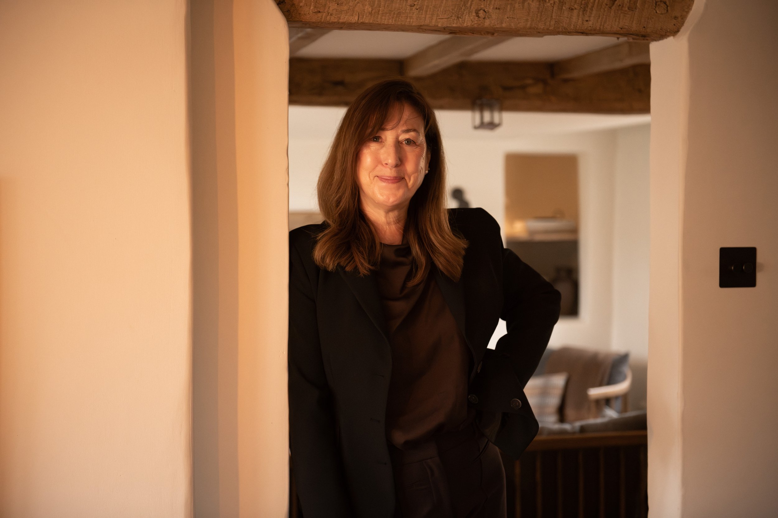 A woman with long brown hair smiling softly, wearing a black blazer, standing indoors behind a wall, with warm lighting and wooden beams visible in the background.