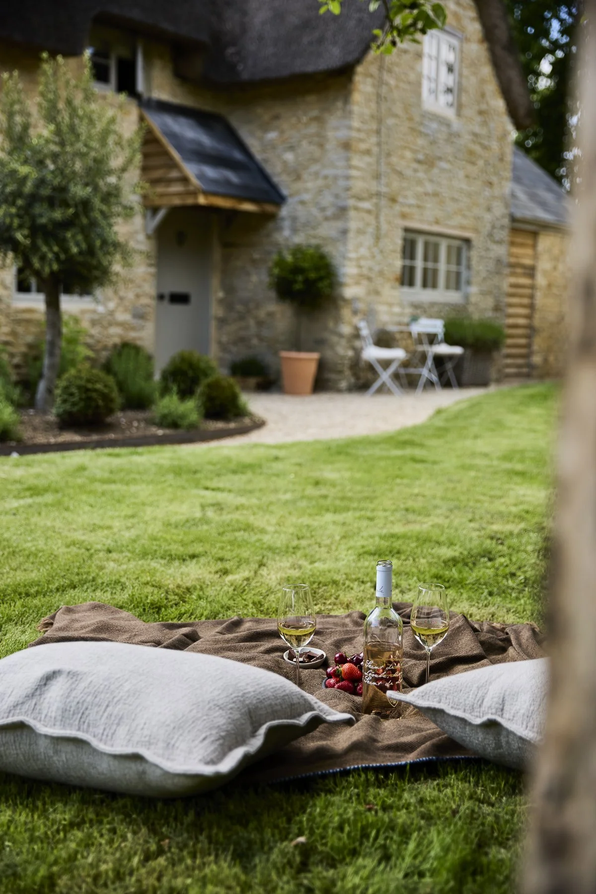 Thatched stone exterior of Nutwood Cottage in Oxfordshire, viewed across a garden lawn with a picnic blanket, cushions and wine glasses.