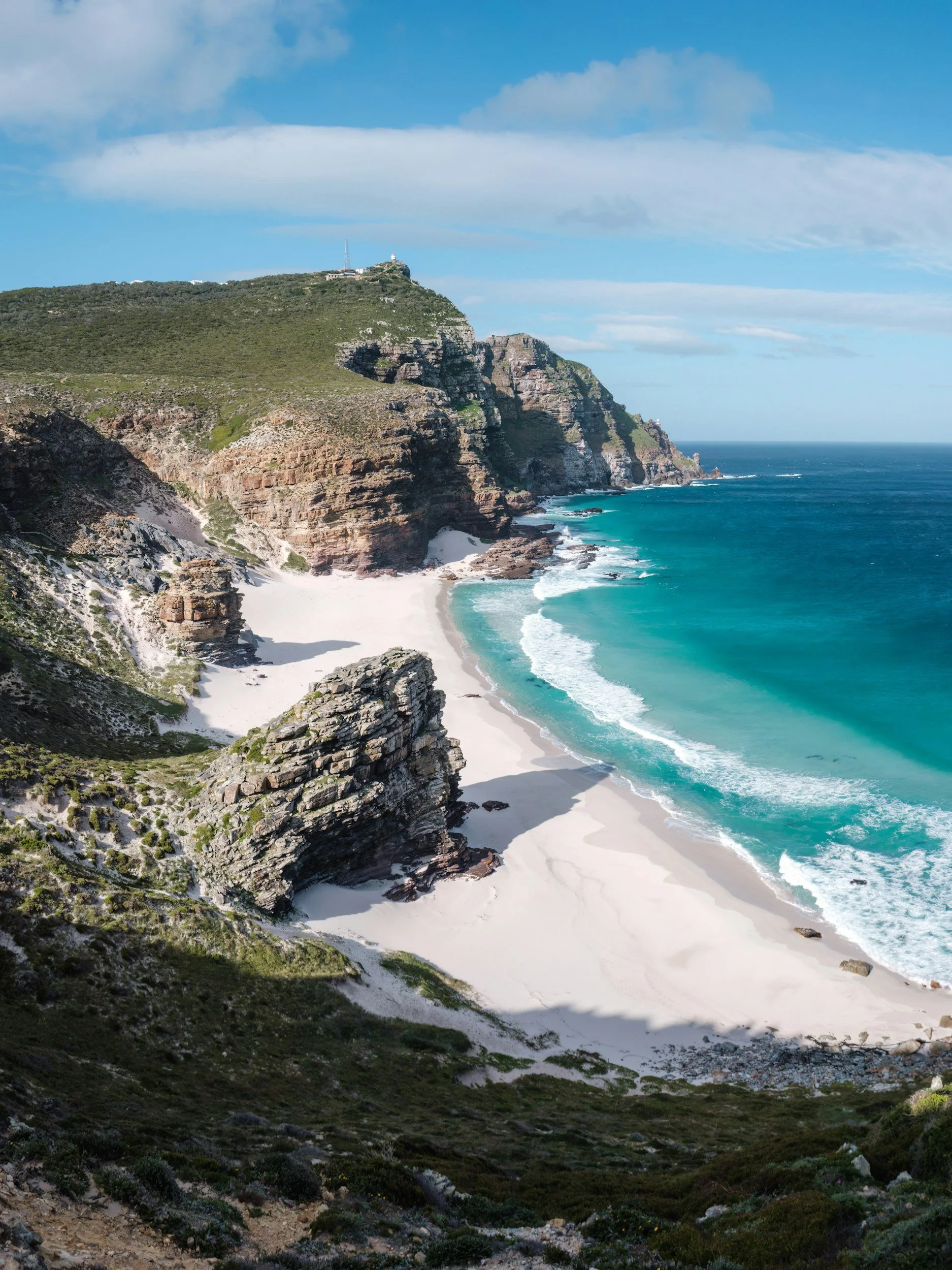 Coastal cliffs with green vegetation, a sandy beach, and blue ocean water under a partly cloudy sky.