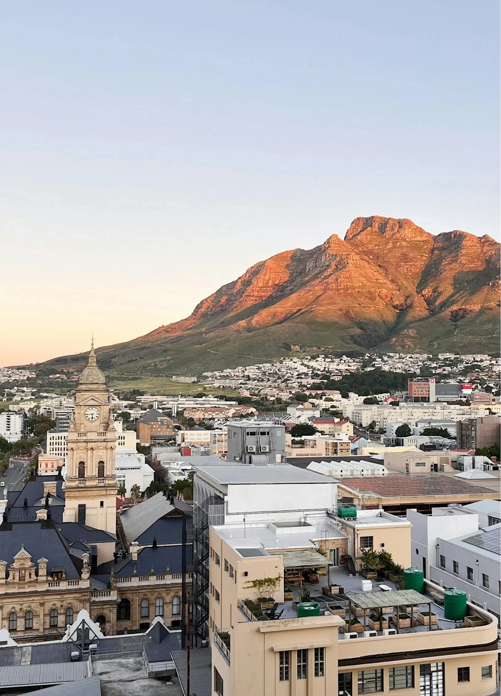 Cityscape with a historic clock tower, modern buildings, and a mountain range in the background during sunset.