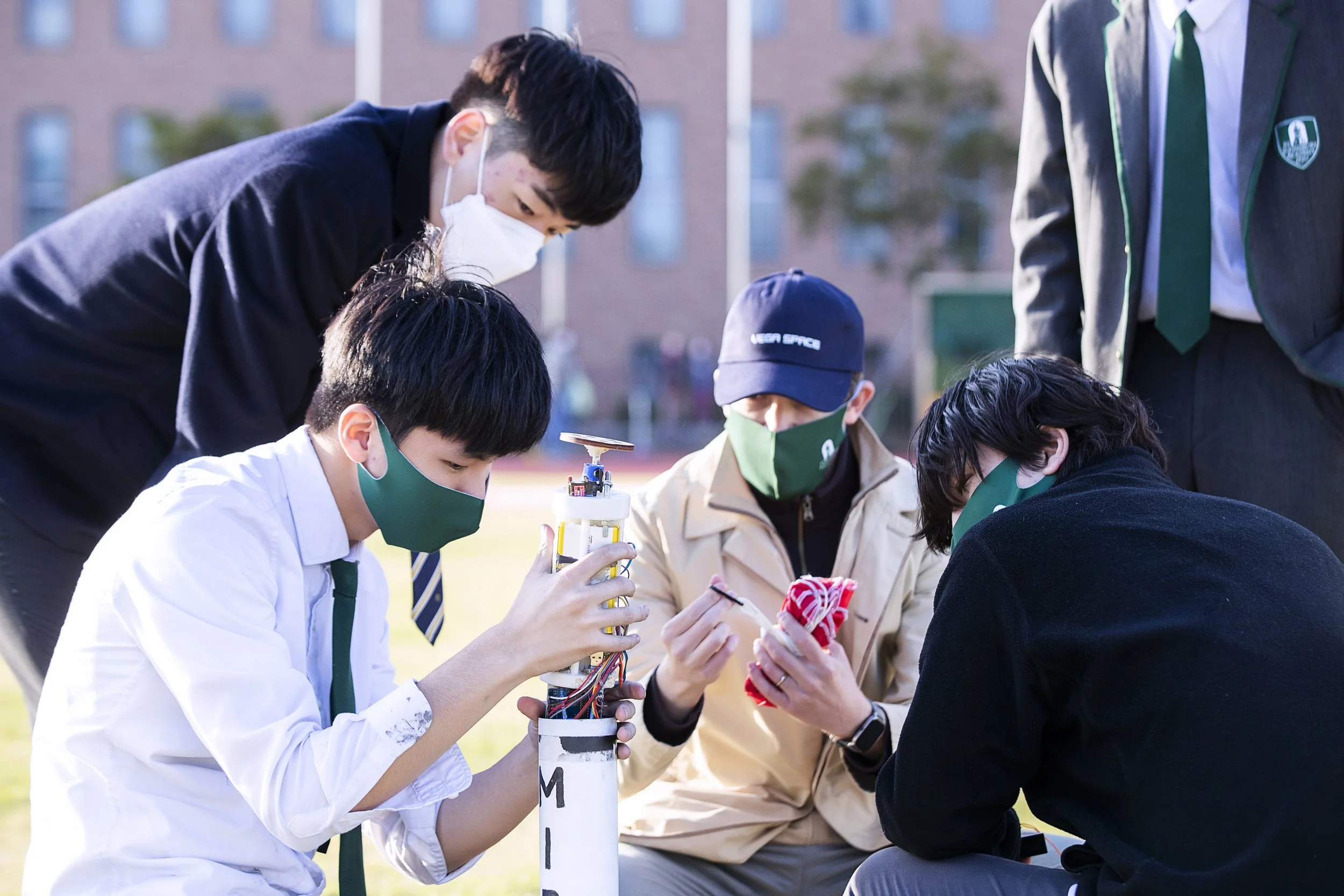 Group of students, wearing masks, gathered around a small rocket-like object outdoors, working on it with tools and a tablet, with a school building in the background.