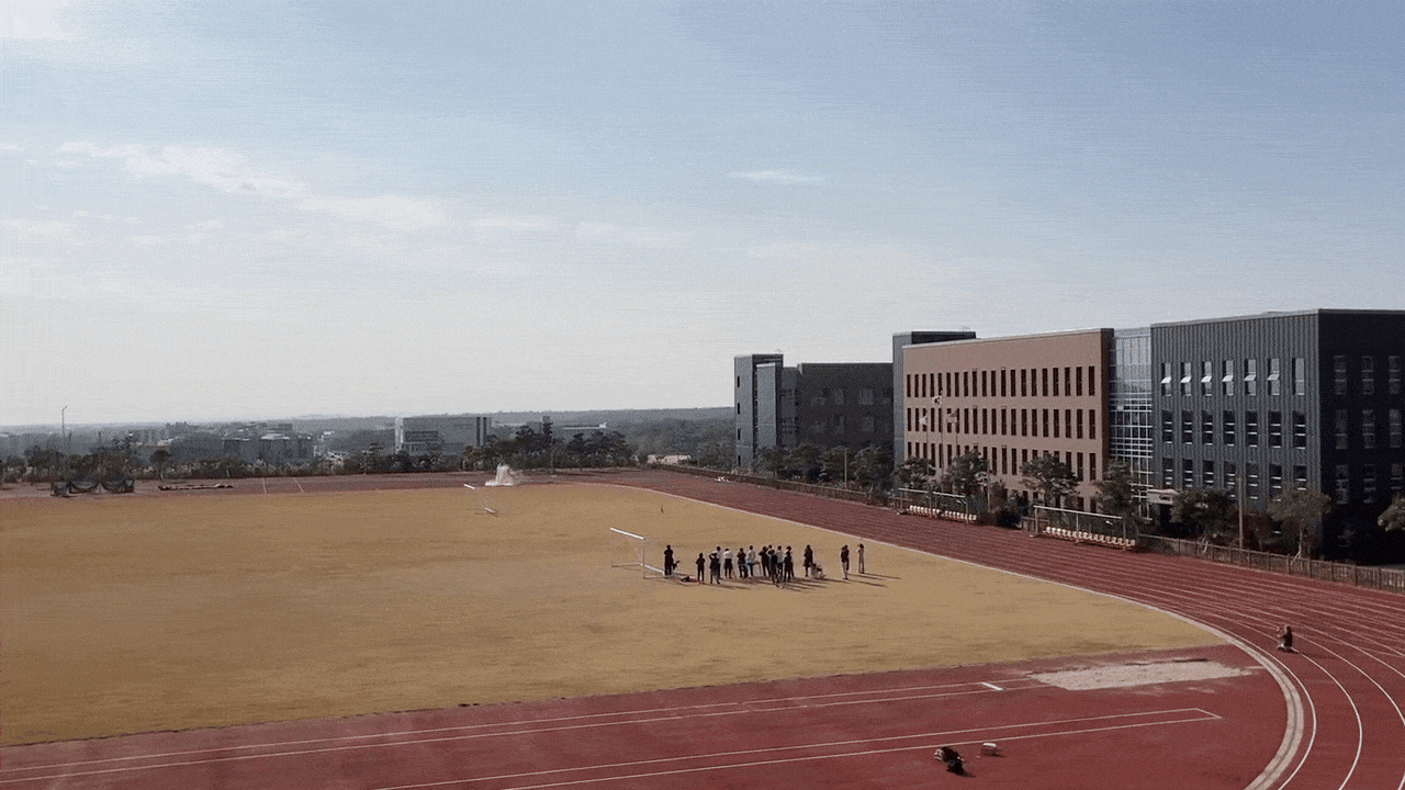 People standing and sitting on a grassy field next to a running track at a school or sports facility, with modern buildings in the background and a clear sky above.