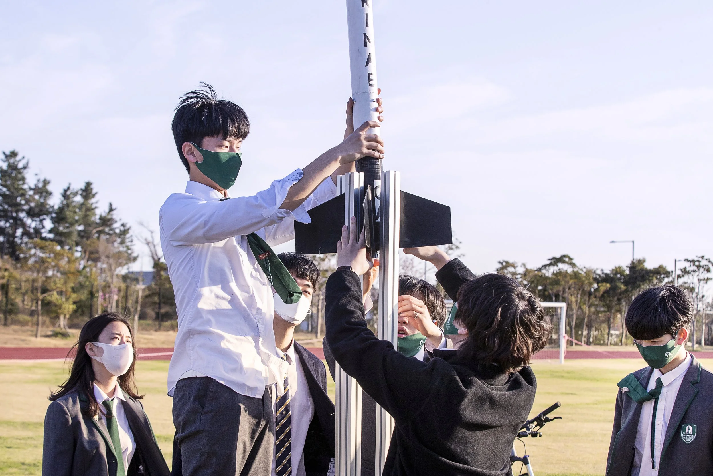 A group of students, wearing school uniforms and face masks, working together on a model rocket outdoors on a school field with trees, track, and soccer goal in the background.
