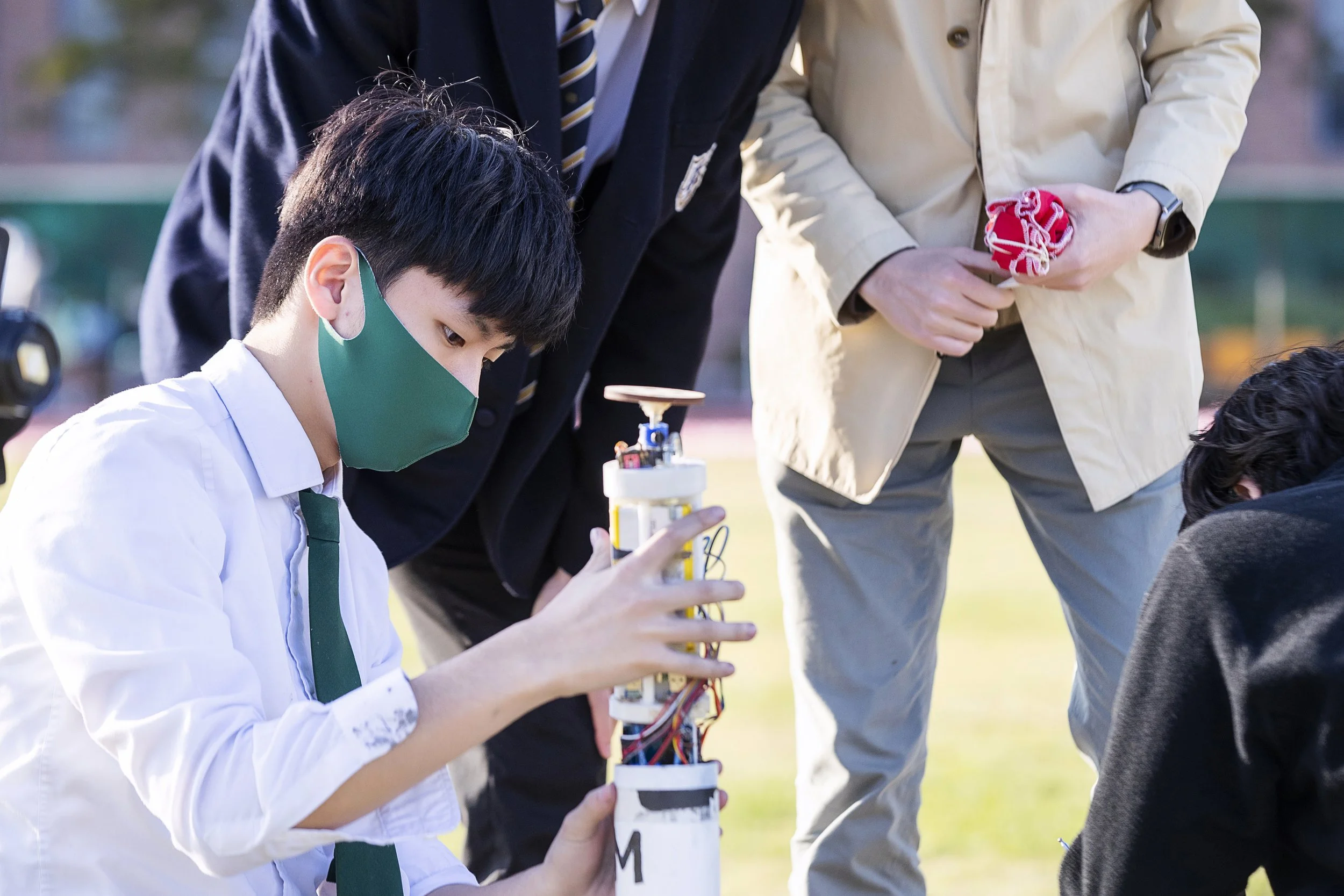 A young boy wearing a green face mask and white shirt with a green tie, holding a small robot with wires and electronic components. Two men are standing nearby, watching him, with one holding a red object. They are outdoors on a grassy area.