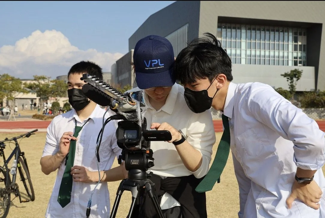 Three students wearing masks and school uniforms, two with green ties and one with a blue cap, are filming or reviewing footage with a video camera outdoors in a school campus.
