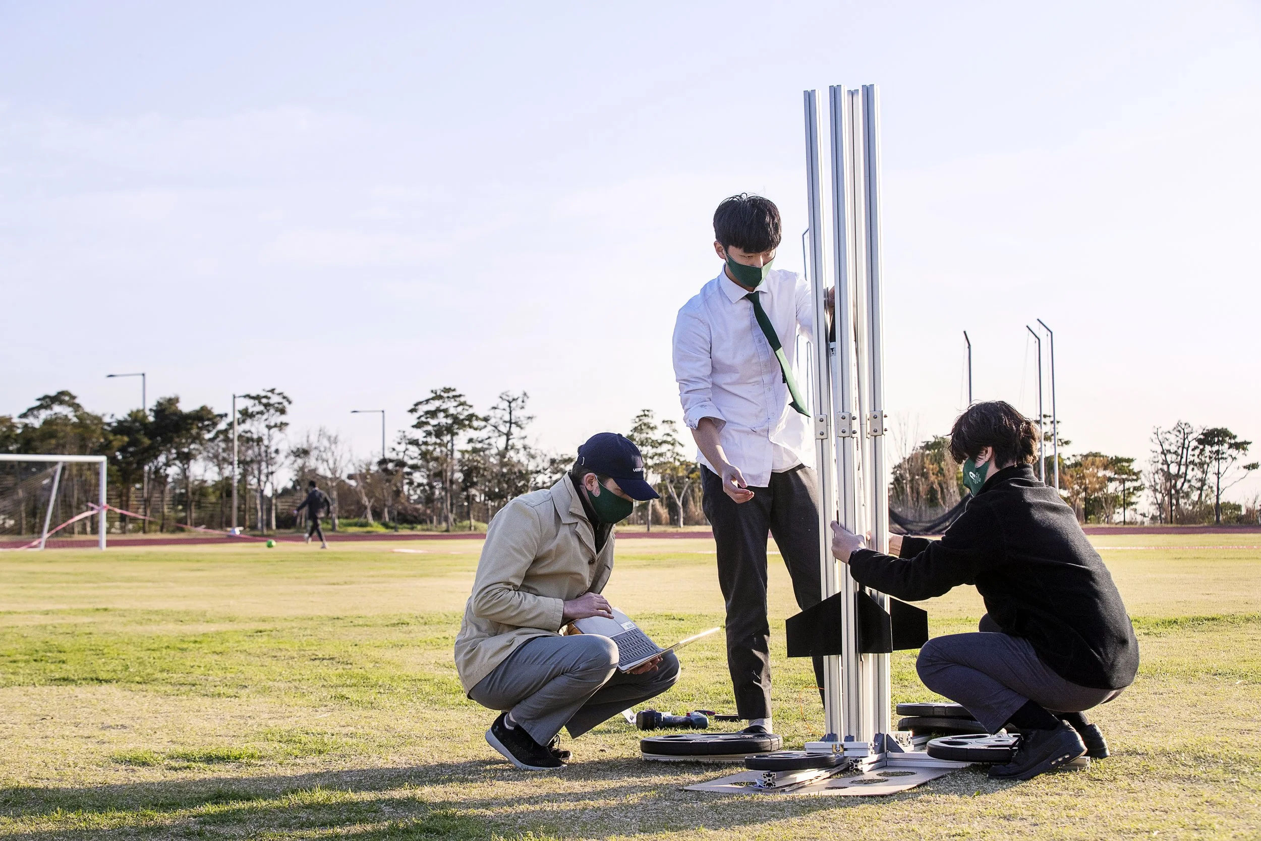 Three individuals assembling a tall metallic structure on a grassy field during daytime, with trees and a running track in the background, all wearing face masks.