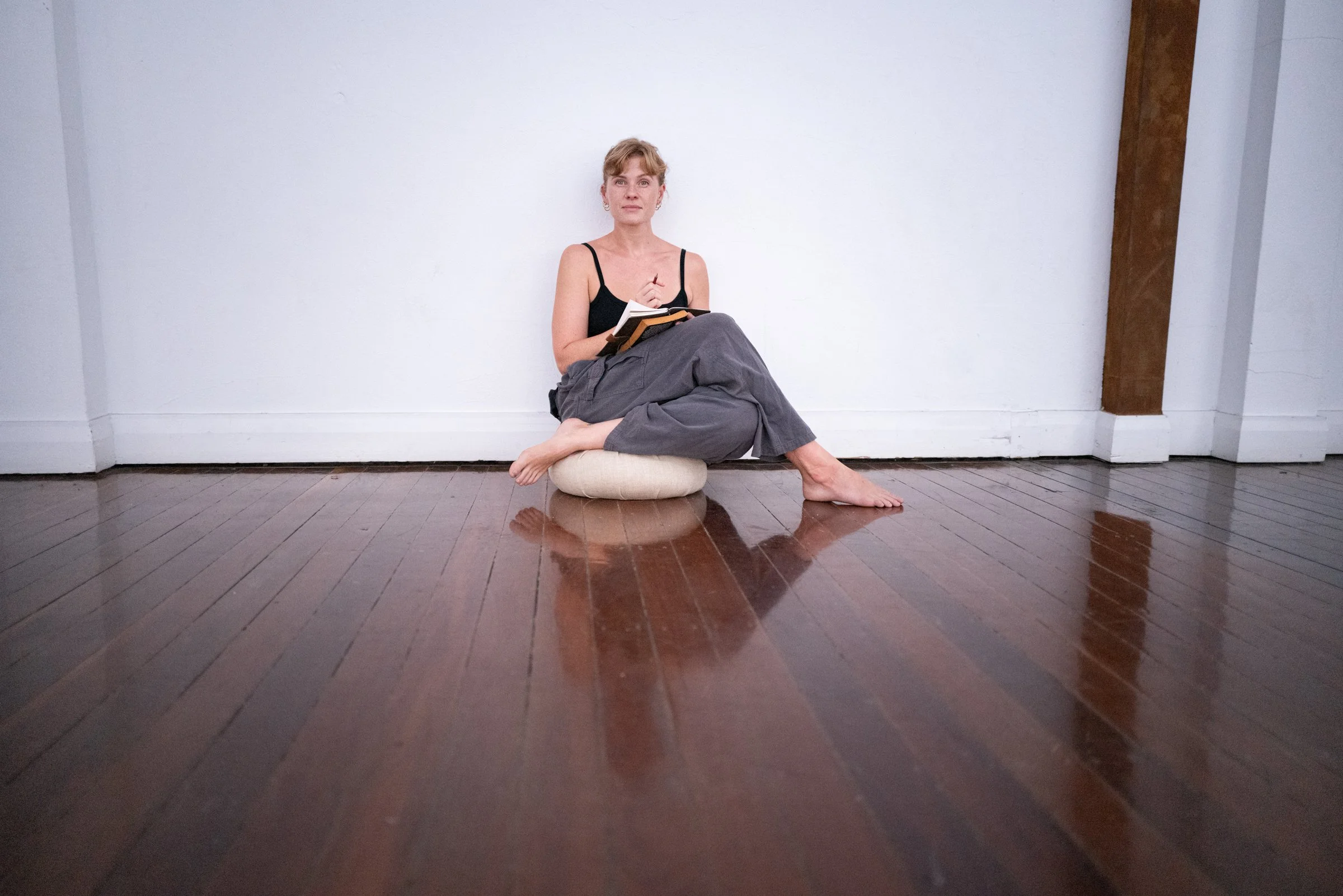 A woman with blonde hair sitting cross-legged on a cushion against a white wall in a room with wooden floors, holding a notepad and pen, wearing a black tank top and gray pants.