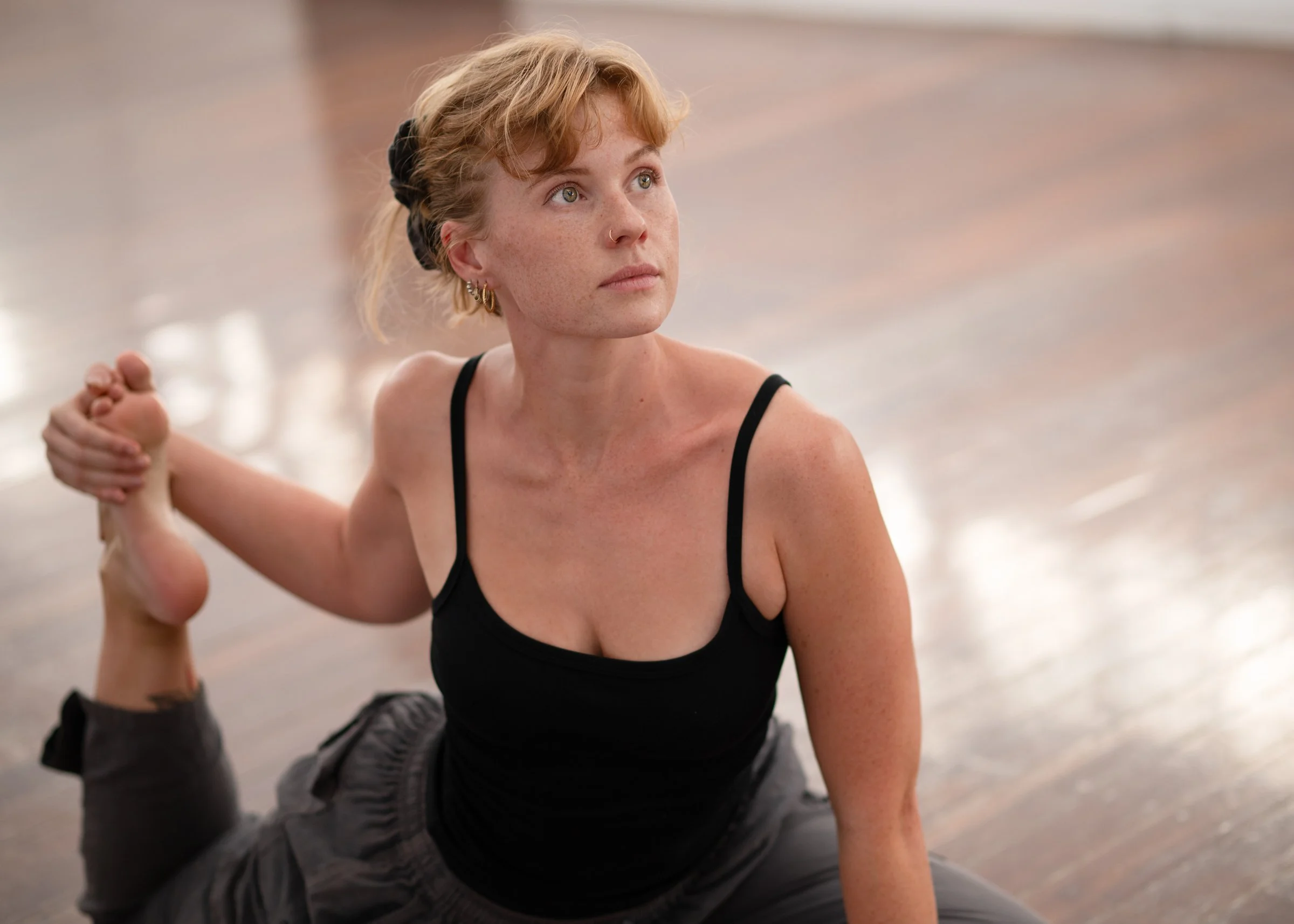 A woman with short wavy red hair and freckles, practicing yoga indoors on a wooden floor. She is wearing a black tank top and dark pants, doing a stretching pose with her right arm holding her foot behind her head.