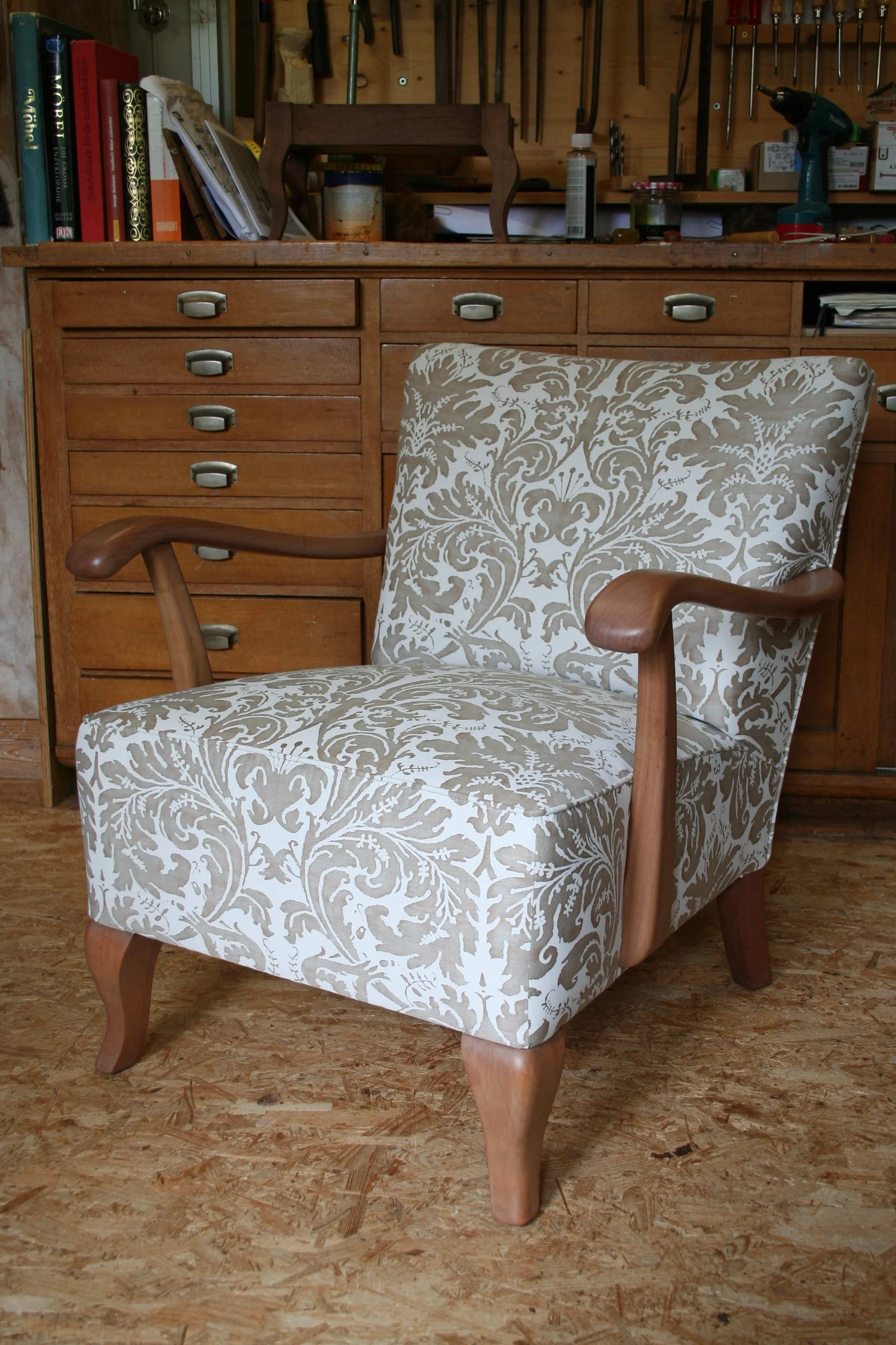 Ein Sessel mit einem weißen Bezug in einem floralem Muster, Holzarmlehnen und -beine, im Hintergrund eine Holzwerkbank mit Werkzeugen und Büchern.