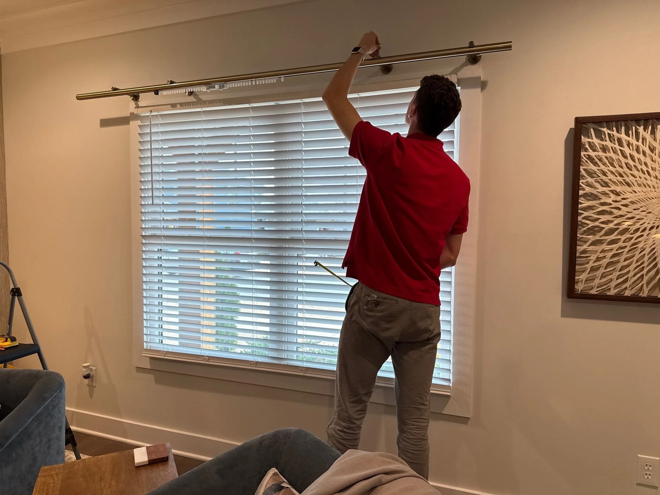 A man in a red shirt is adjusting a curtain rod in a room with beige walls and white window blinds.