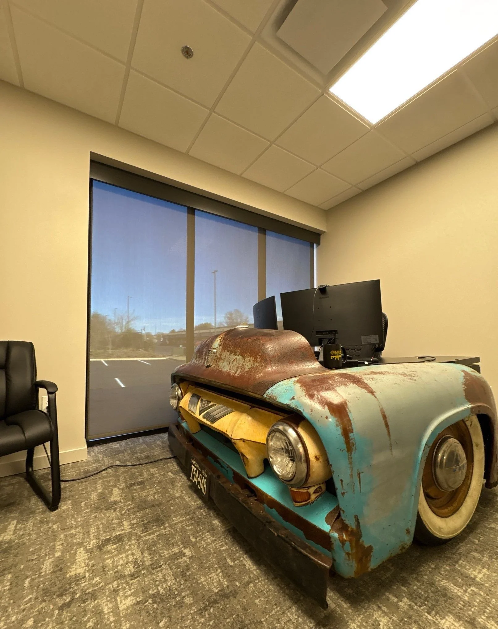 An office with a vintage car-shaped desk, black office chair, window, computer monitor, and ceiling lights.