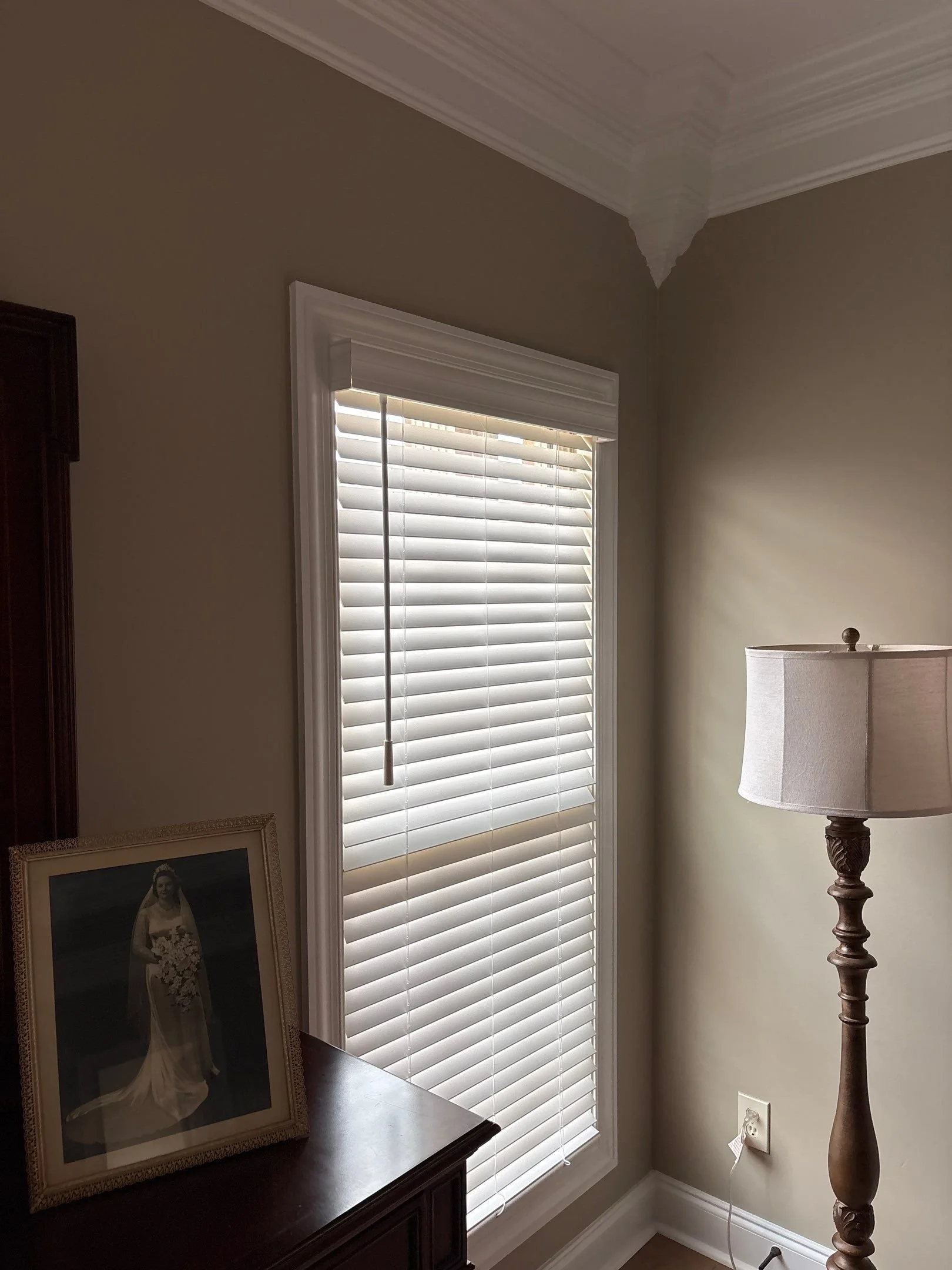 A corner of a room with a window covered by white blinds, a wooden dresser with a framed photo of a woman in a wedding dress, a standing lamp with a beige shade, beige walls, a white electrical outlet, and crown molding on the ceiling.