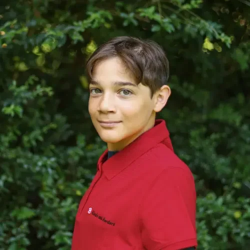 A young boy with short brown hair and tan skin, wearing a red polo shirt, standing outdoors in front of green foliage.