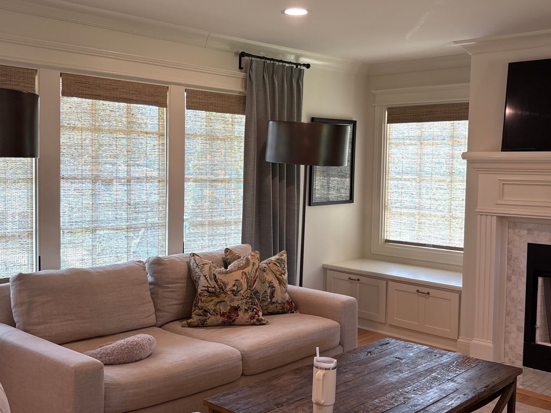 Living room with beige sofa, floral pillows, large windows with woven shades, and a white fireplace mantel. A black floor lamp and a framed picture are on the walls. A wooden coffee table is in front of the sofa with a white candle and a hypoallergenic cover on the armrest.