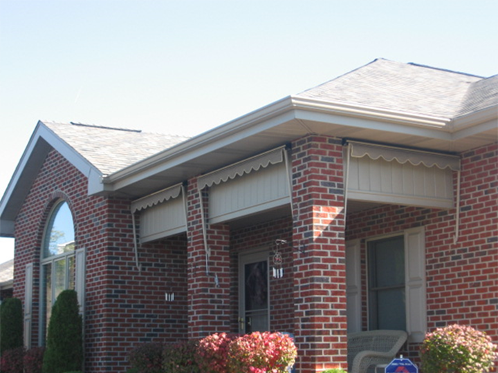 Front view of a red brick house with two window awnings, a glass door, and a small porch with bushes and plants.