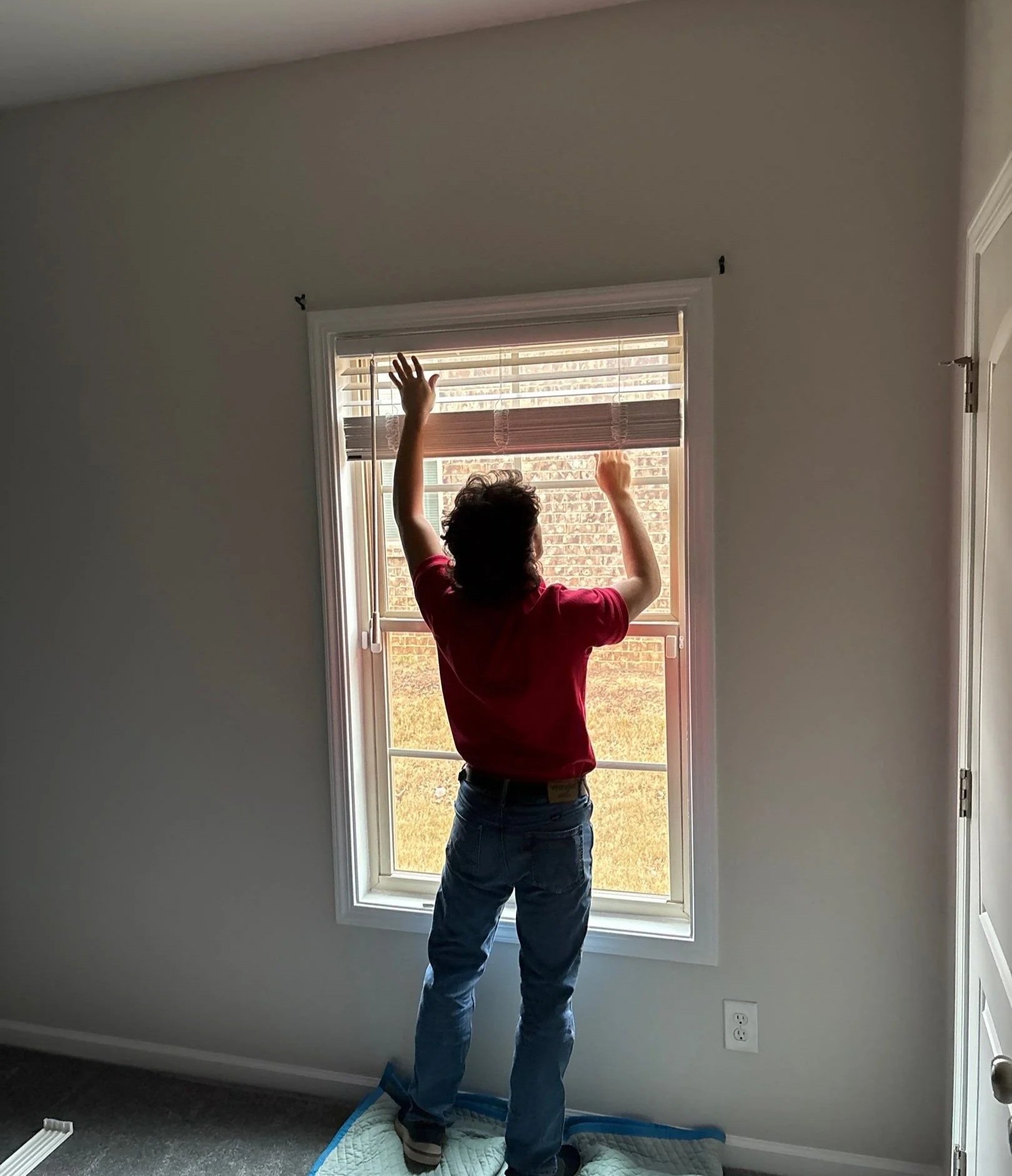 A person standing on a mattress on the floor, adjusting blinds on a window in a minimally decorated room with light gray walls and dark carpet flooring.