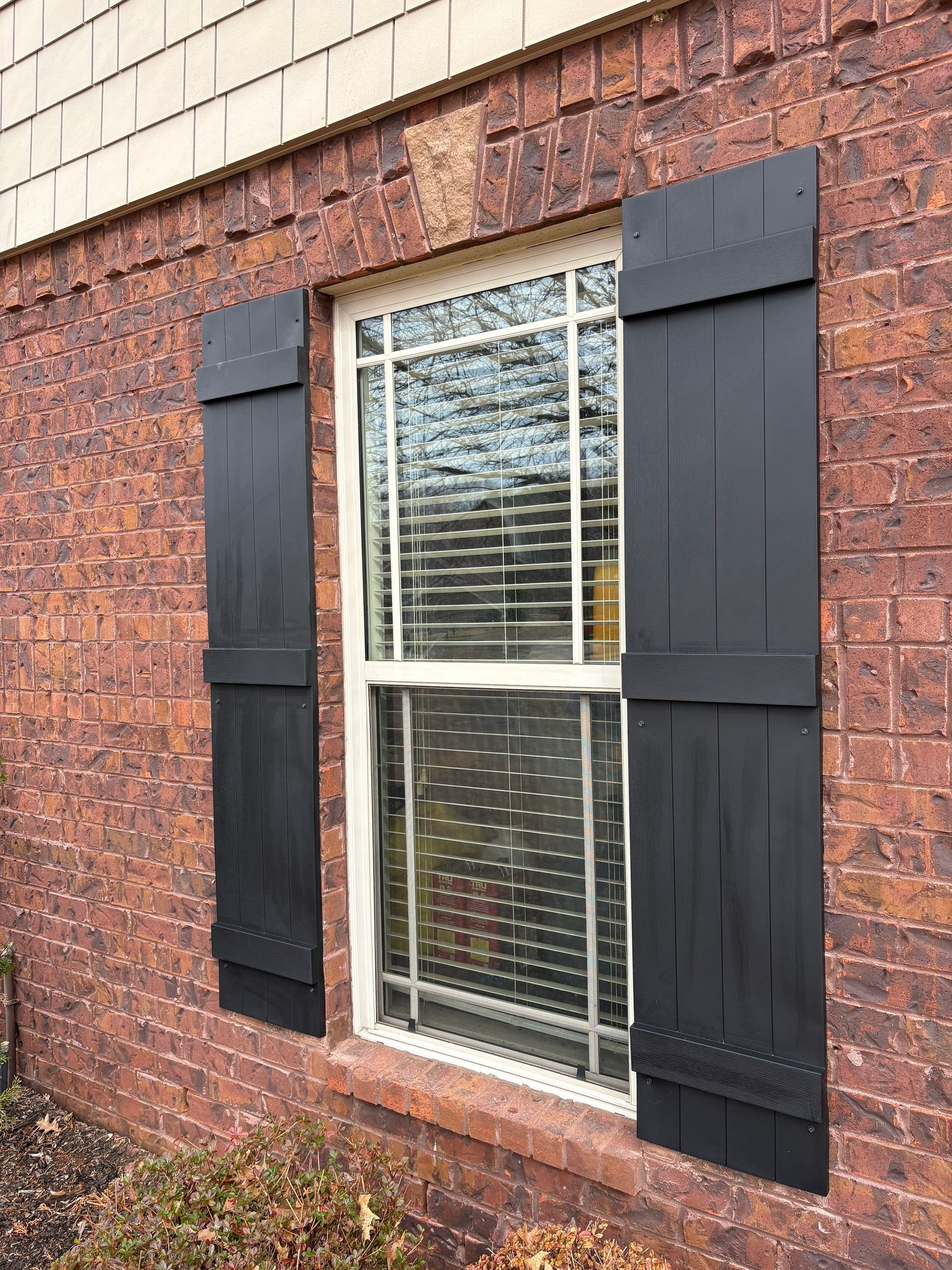 A red brick house wall with a white-framed window and black decorative shutters on either side. Outside, there are small plants at the base of the wall and some fallen leaves on the ground.