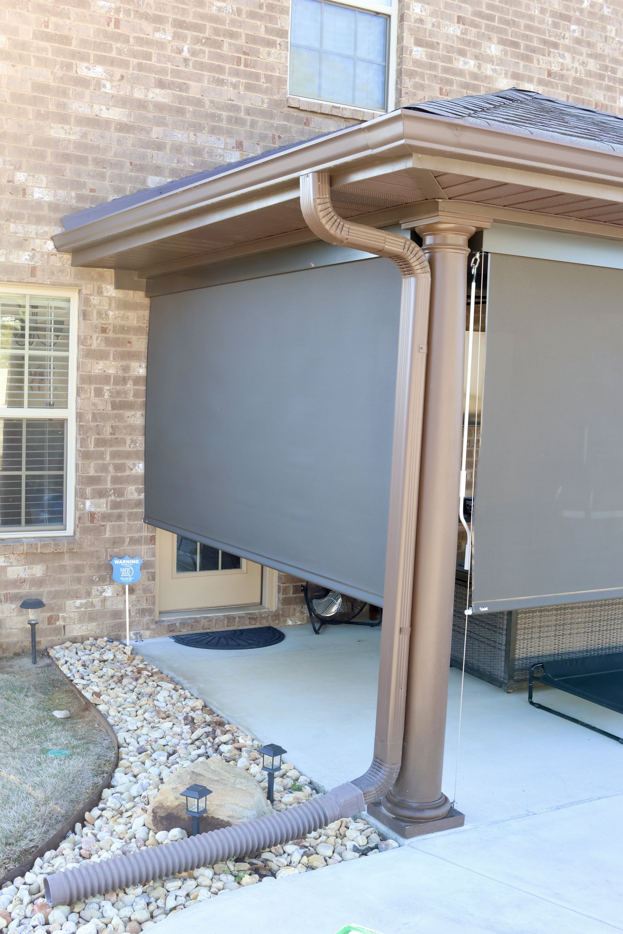 Exterior view of a house with a brick wall, two windows, a door with a decorative mat, and a small landscaped area with rocks, small lamps, and a brown installation with a gutter system and a retractable sunshade.