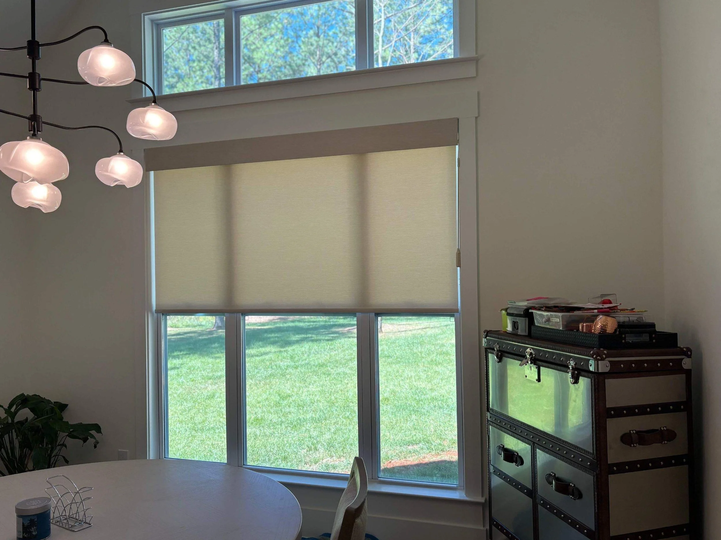 A bright room with an oval dining table, partially visible, with a small houseplant and a container on it. There is a large window with a beige roller blind, showing a green lawn and trees outside. A three-light chandelier with pinkish glass shades hangs from the ceiling. To the right, there is a black metal and wood storage cabinet with books and various items on top.
