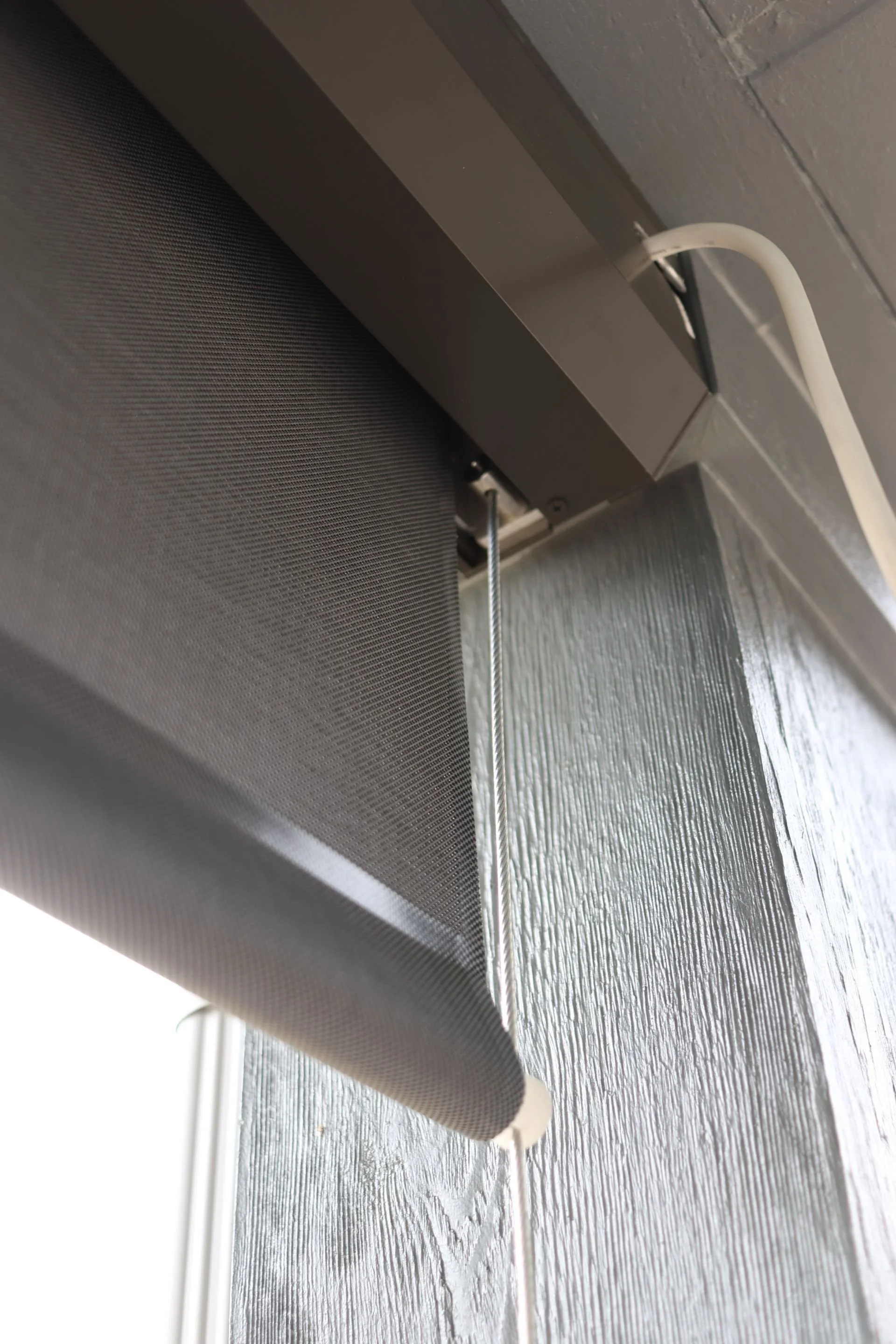 Close-up of a black roller shade window covering with a white electrical cord attached, mounted on a gray textured wall.