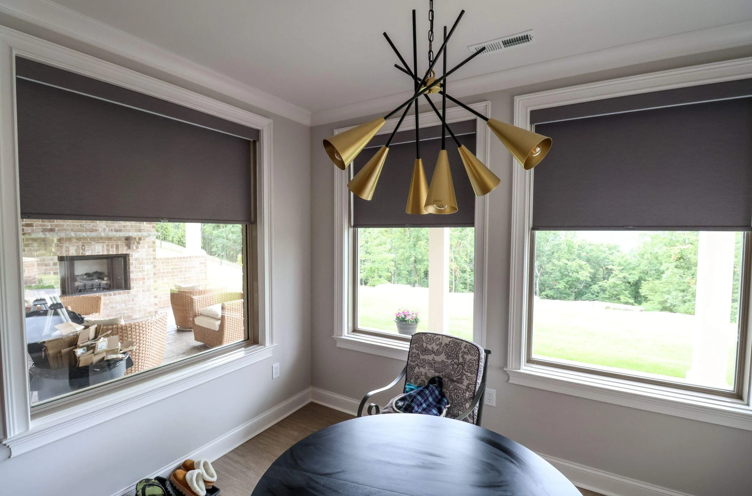 Interior view of a room with large windows and a modern black and gold chandelier hanging from the ceiling. Outside, there are trees and a lawn visible through the windows. Inside, a chair with a patterned cushion and a round black table are visible, along with some shoes on the floor and a glimpse of a patio area with wicker chairs and a brick fireplace.