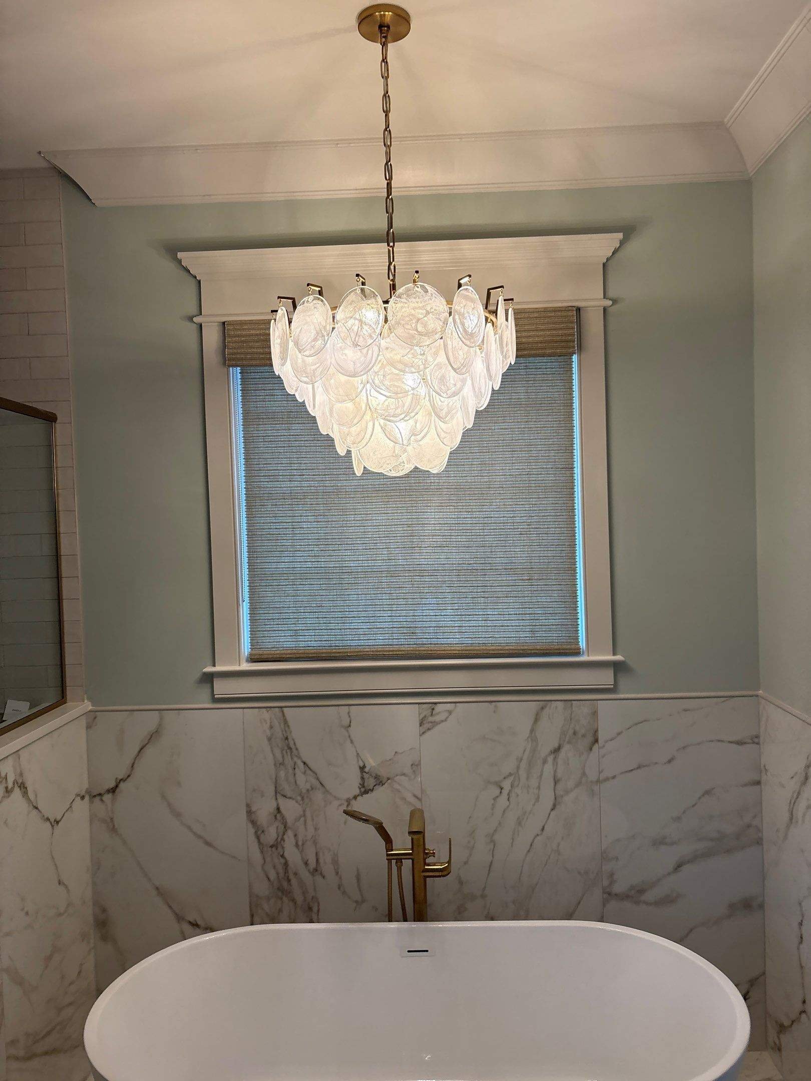 Modern bathroom with bathtub, gold faucet, marble walls, window with blinds, and a chandelier hanging from the ceiling.